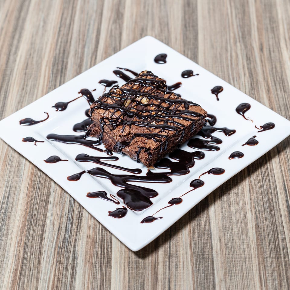 A square chocolate brownie drizzled with chocolate sauce on a white square plate.